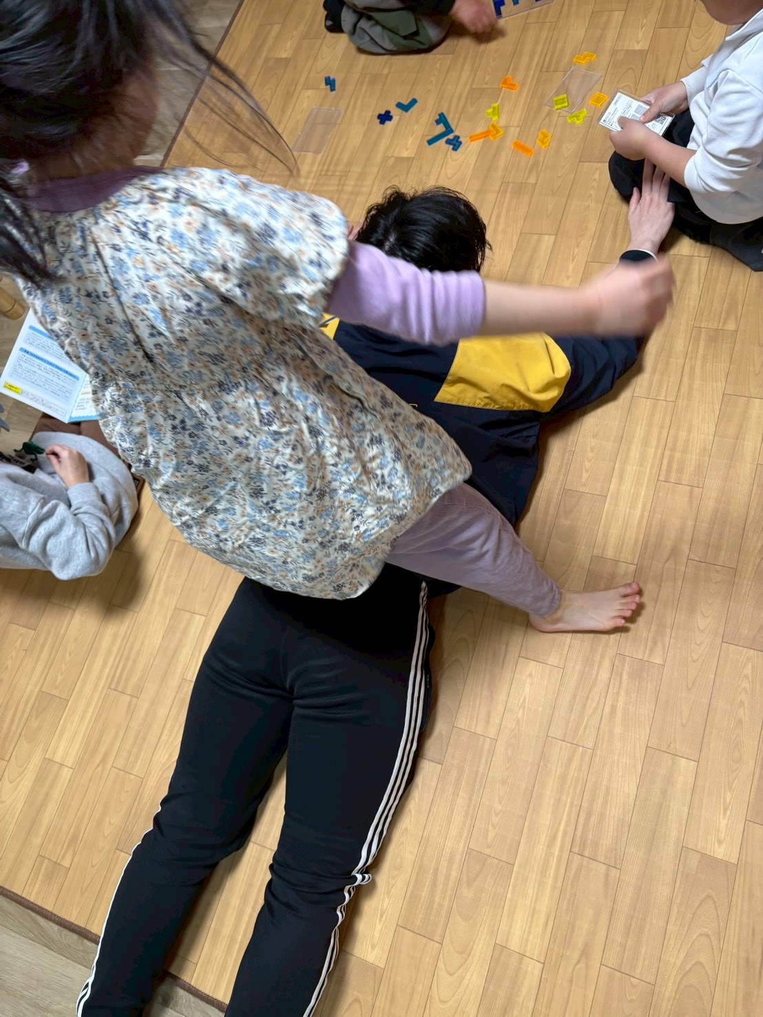 「鉄壁の亀（スタッフ）。」床に張り付いて「絶対動かないぞ！」と踏ん張るスタッフの背中と、作戦を練る子どもたち。