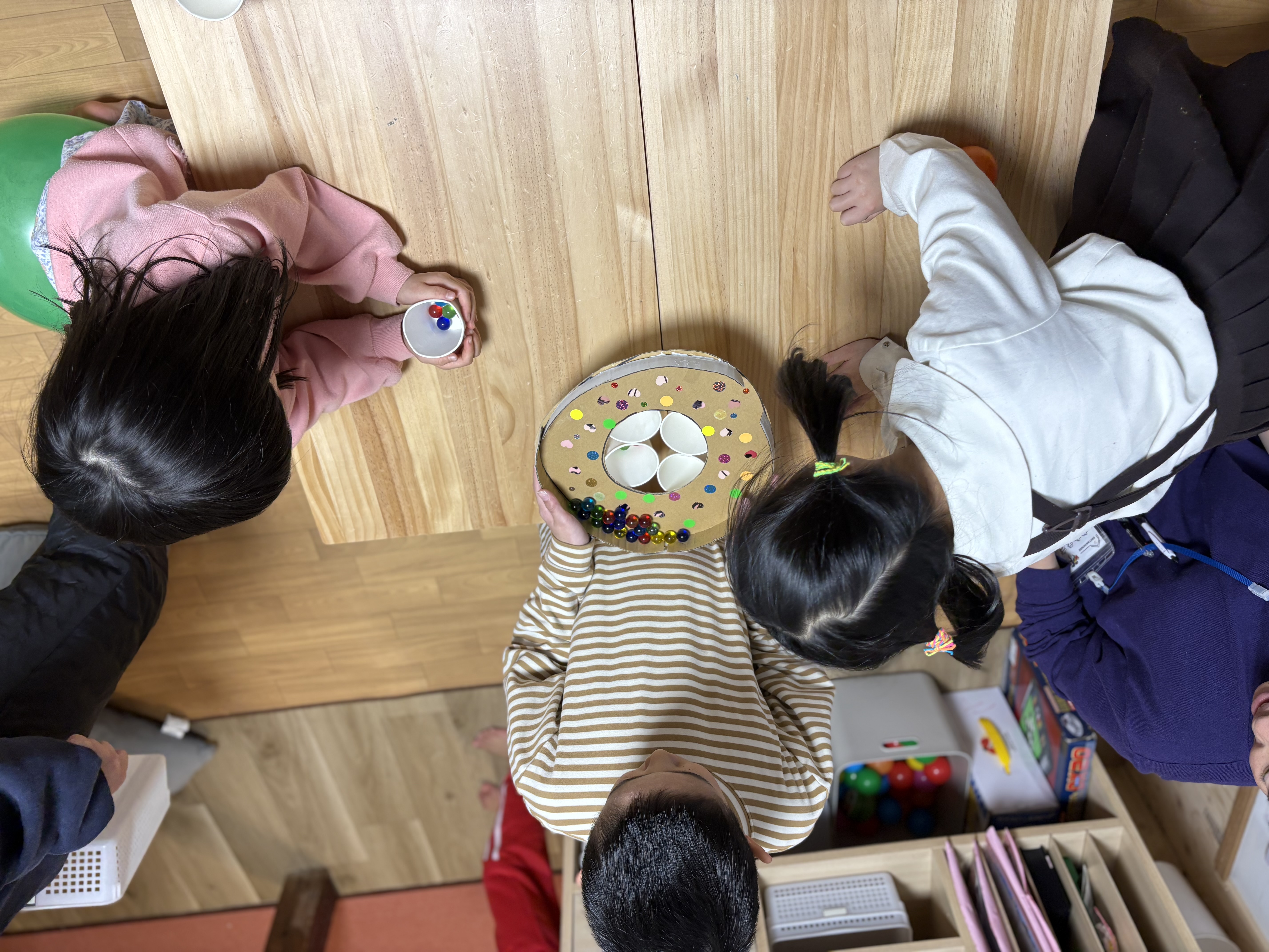 がんばれー！の全力応援。」

友達がビー玉を落とす姿を、身を乗り出して応援する子どもたちの活気ある表情。