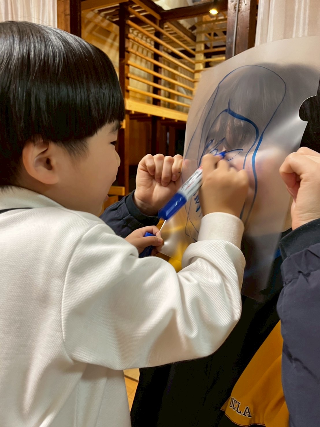 「先生の顔、貸してね！」

クリアファイルを顔に当てたスタッフと、ペンを握って「どこから描こうかな？」と狙いを定めるお子様の、遊び心溢れる対面ショット。