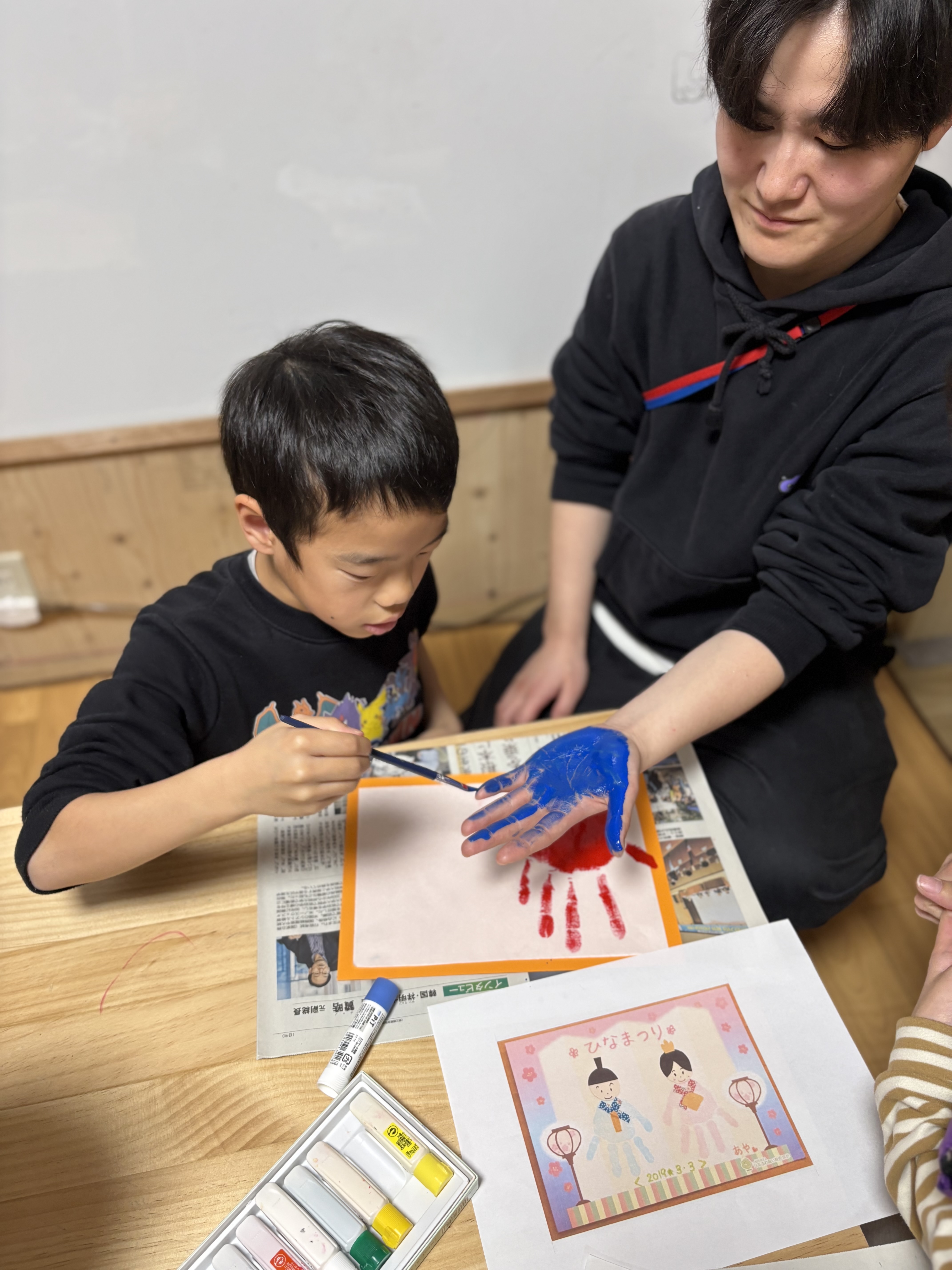 「筆先の、くすぐったいコミュニケーション。」
スタッフの手のひらに、慎重に筆を滑らせる瞬間。塗られるスタッフの「わはは!」という笑顔と、塗るお子様の「真剣なパティシエ」のような対照的なカット。