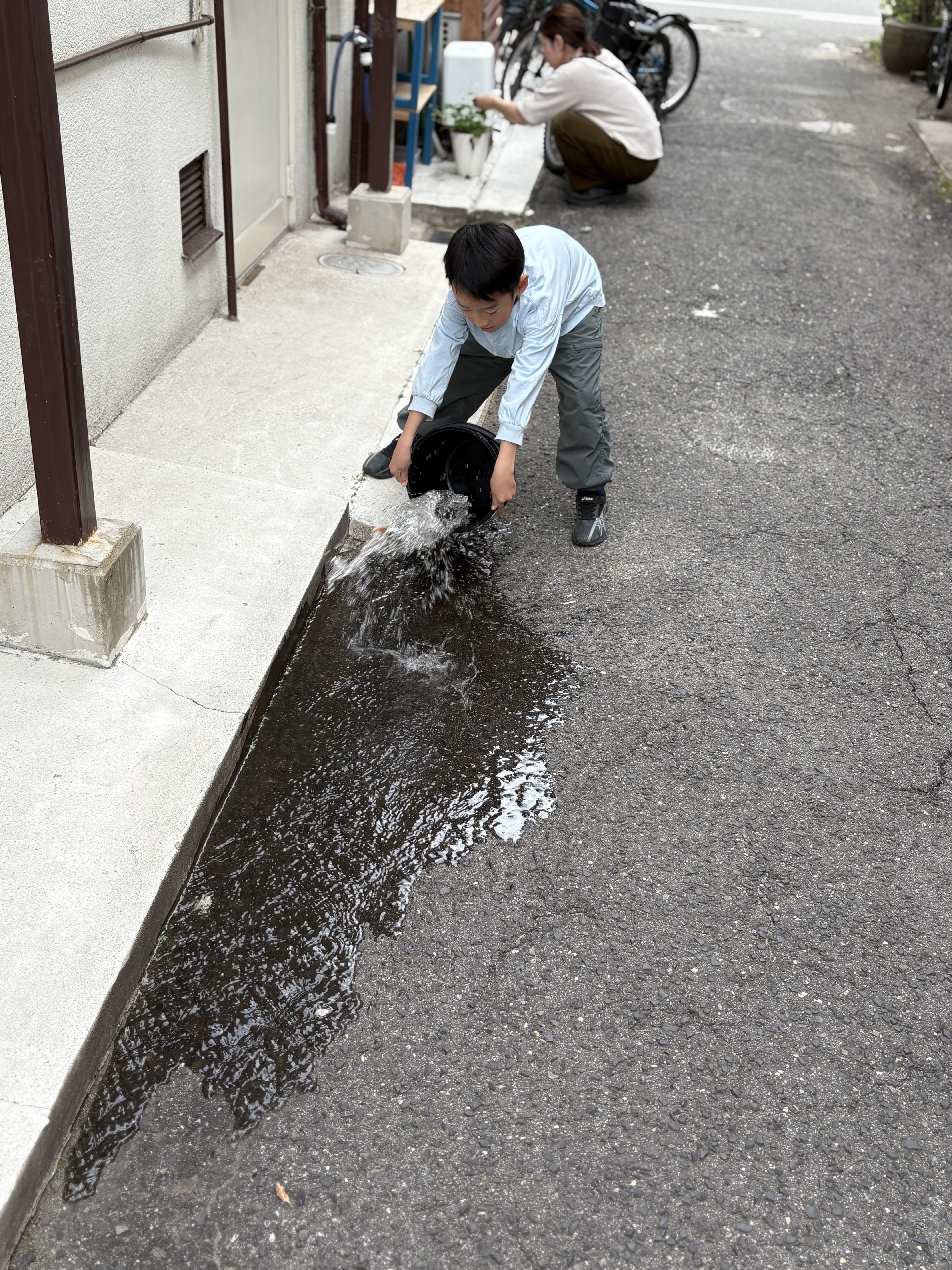 「一気に流す！砂埃よ、さようなら。」

バケツの水を地面に勢いよく撒くダイナミックな瞬間。水しぶきが舞い、地面の色がパッと変わる躍動感のある1枚。