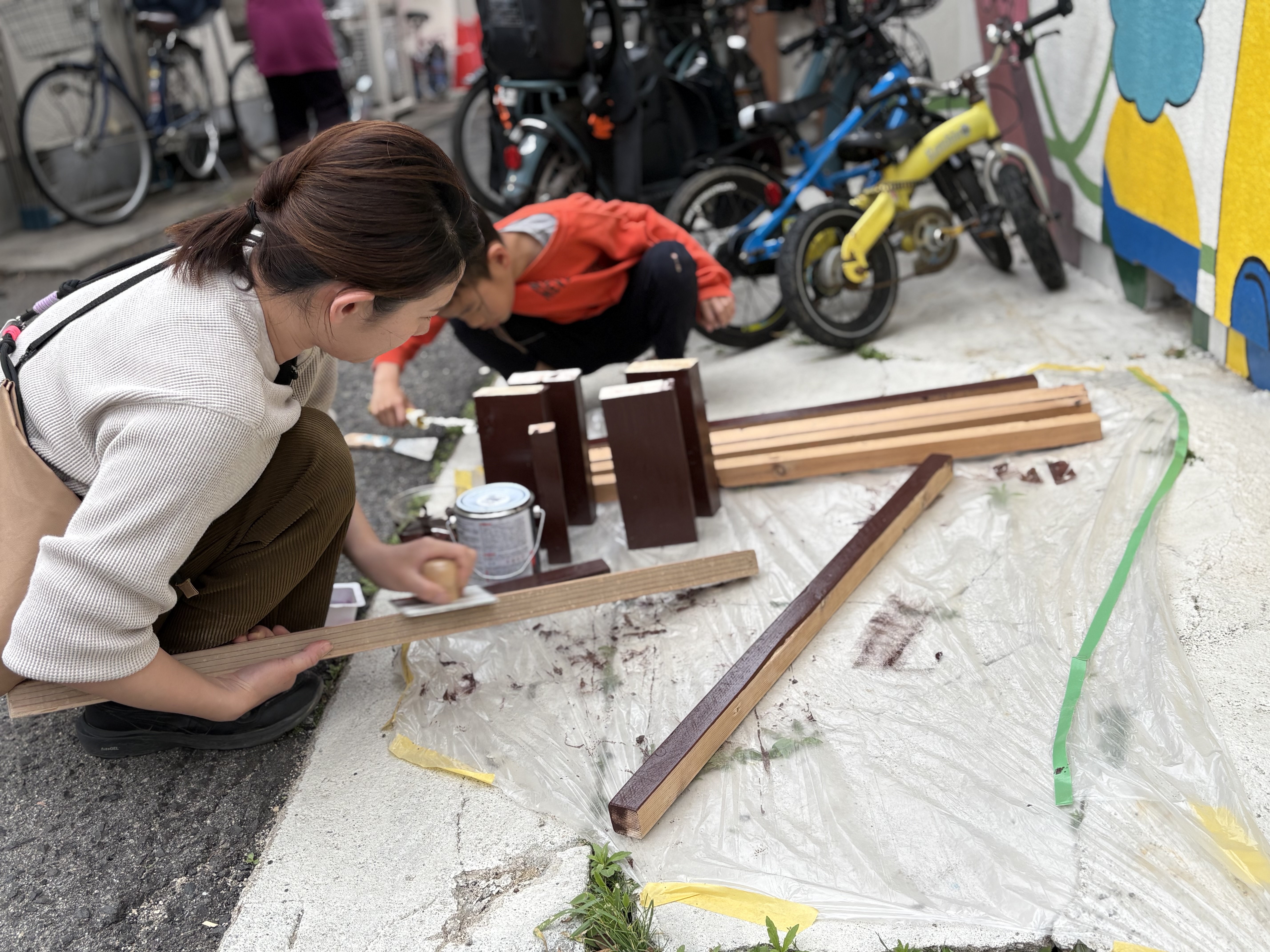 「一塗りずつ、温かみを乗せて。」

ベンチの木目に沿って、なおちゃんと並んでペンキを塗る手元。共同作業の「絆」を感じさせるカット。