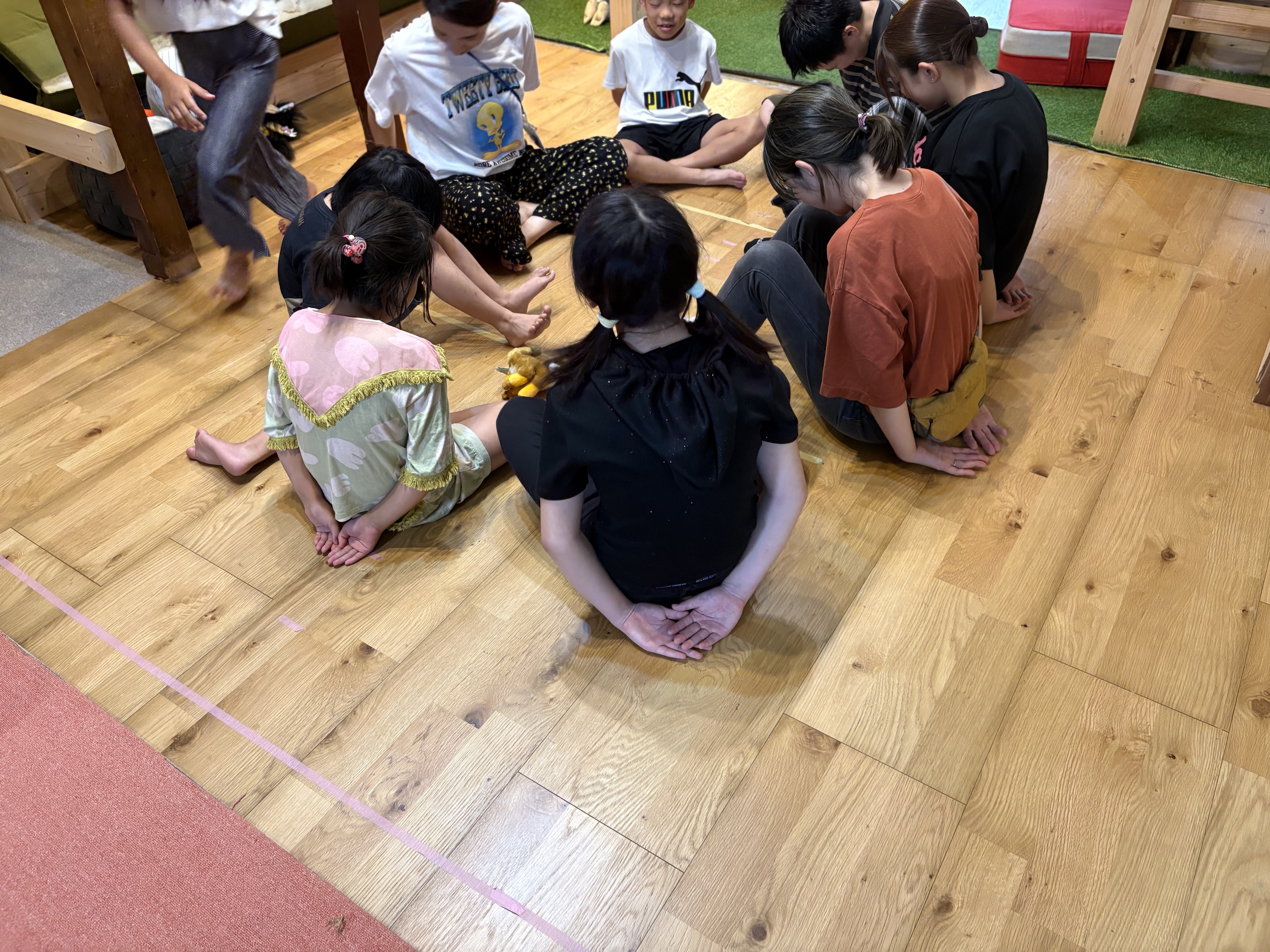「作戦会議室。円になって床に座り、いざ準備完了!」
子どもたちとスタッフがきれいな円を作って床に座り、後ろに手を組んで「誰に落とされるかな…」とワクワクが始まる引きショット。