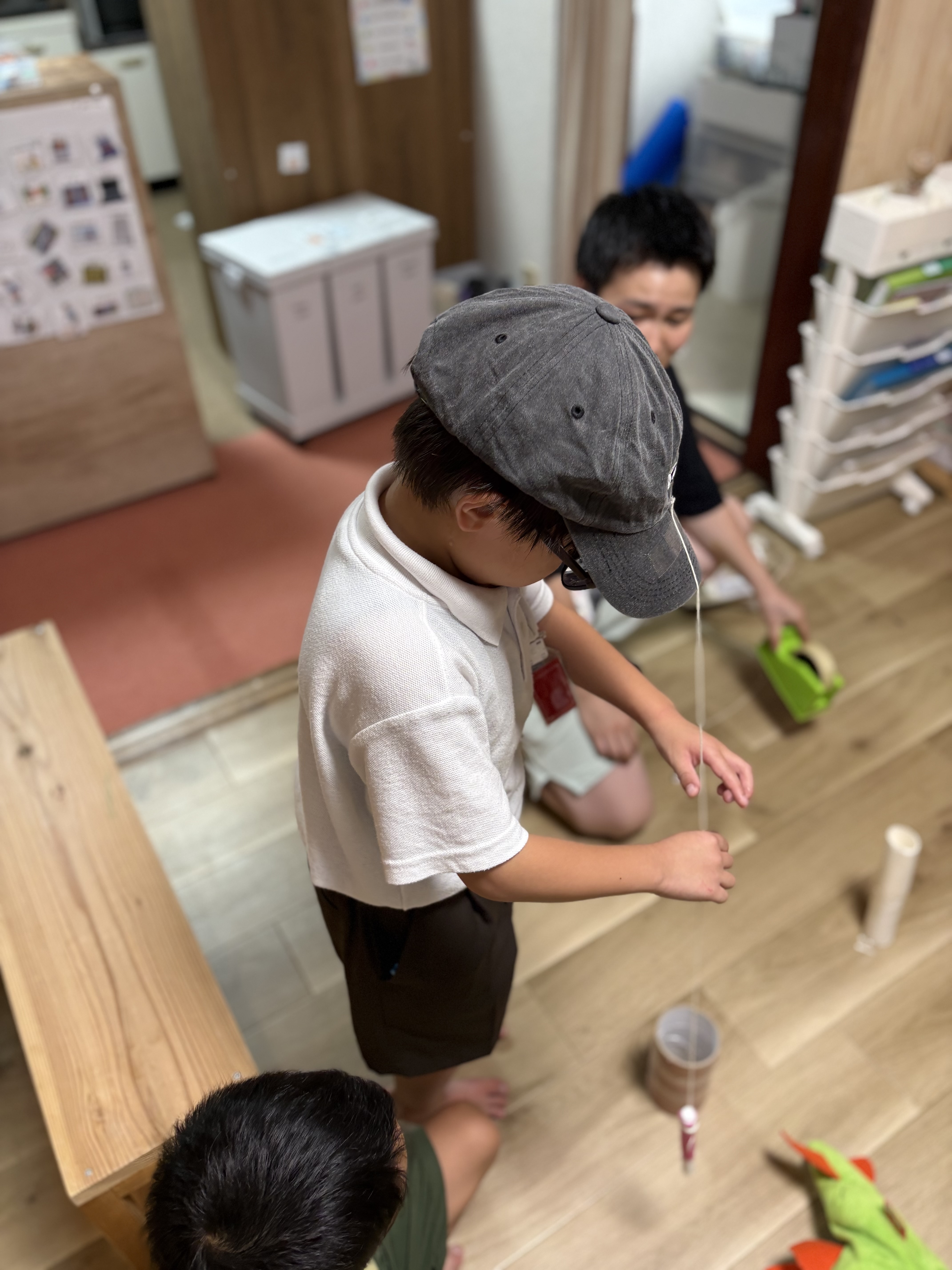 「本日開幕！謎の特製『ハットインゲーム』に興味津々。」

ひもとペンがついた不思議な帽子を前に、「これどうやって遊ぶの！？」とワクワクしながらスタッフの説明をきく子どもたちの引きショット。