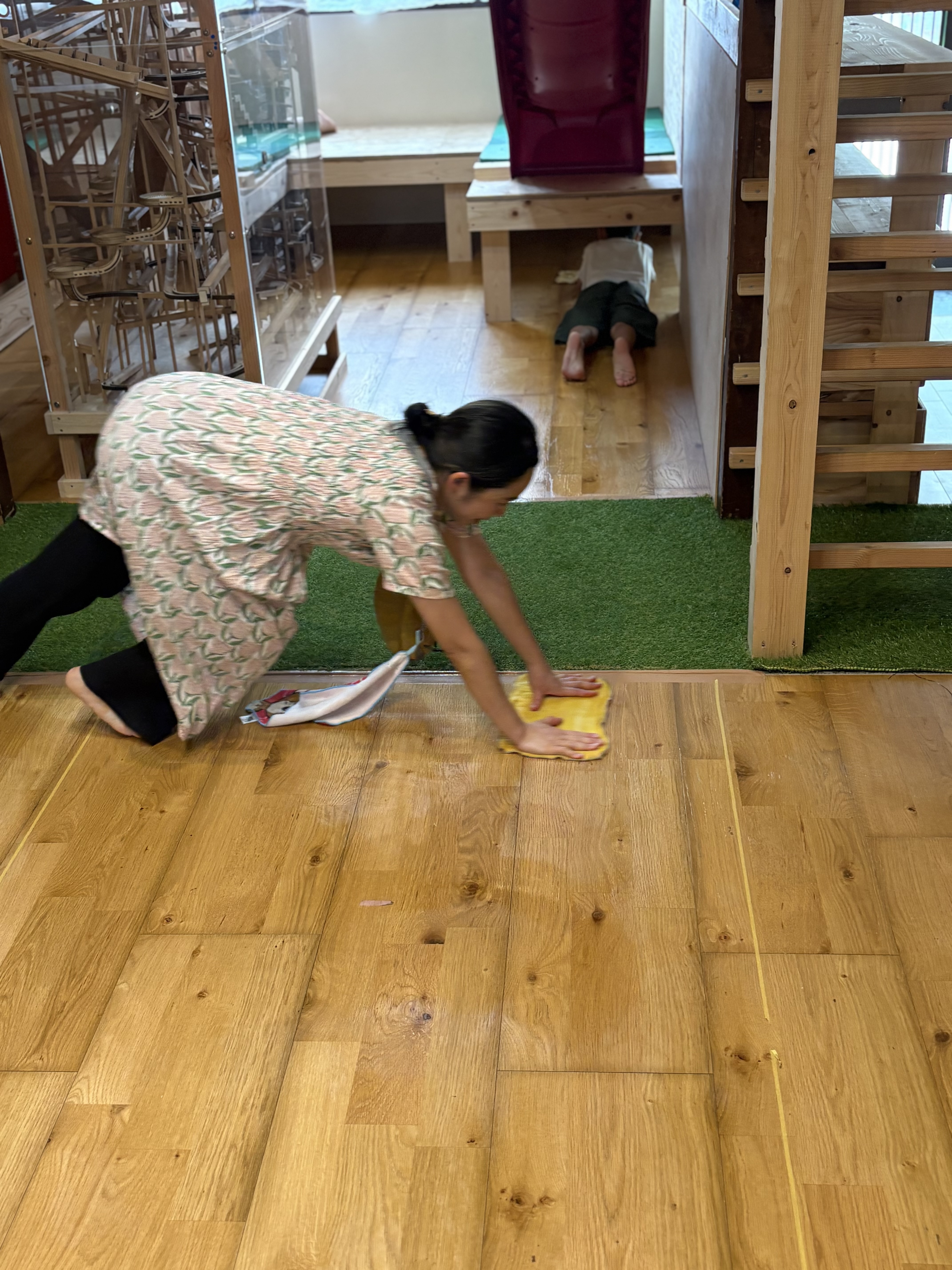 「『先生、そこまだ汚れてんで!』子ども監督から厳しいチェックが入る瞬間」
スタッフが木工遊具の脚の周りを一生懸命にゴシゴシと拭いている姿を、横から子どもが「そこそこ!がんばれー!」と嬉しそうに指差して応援(監督)している微笑ましいひとコマ。
