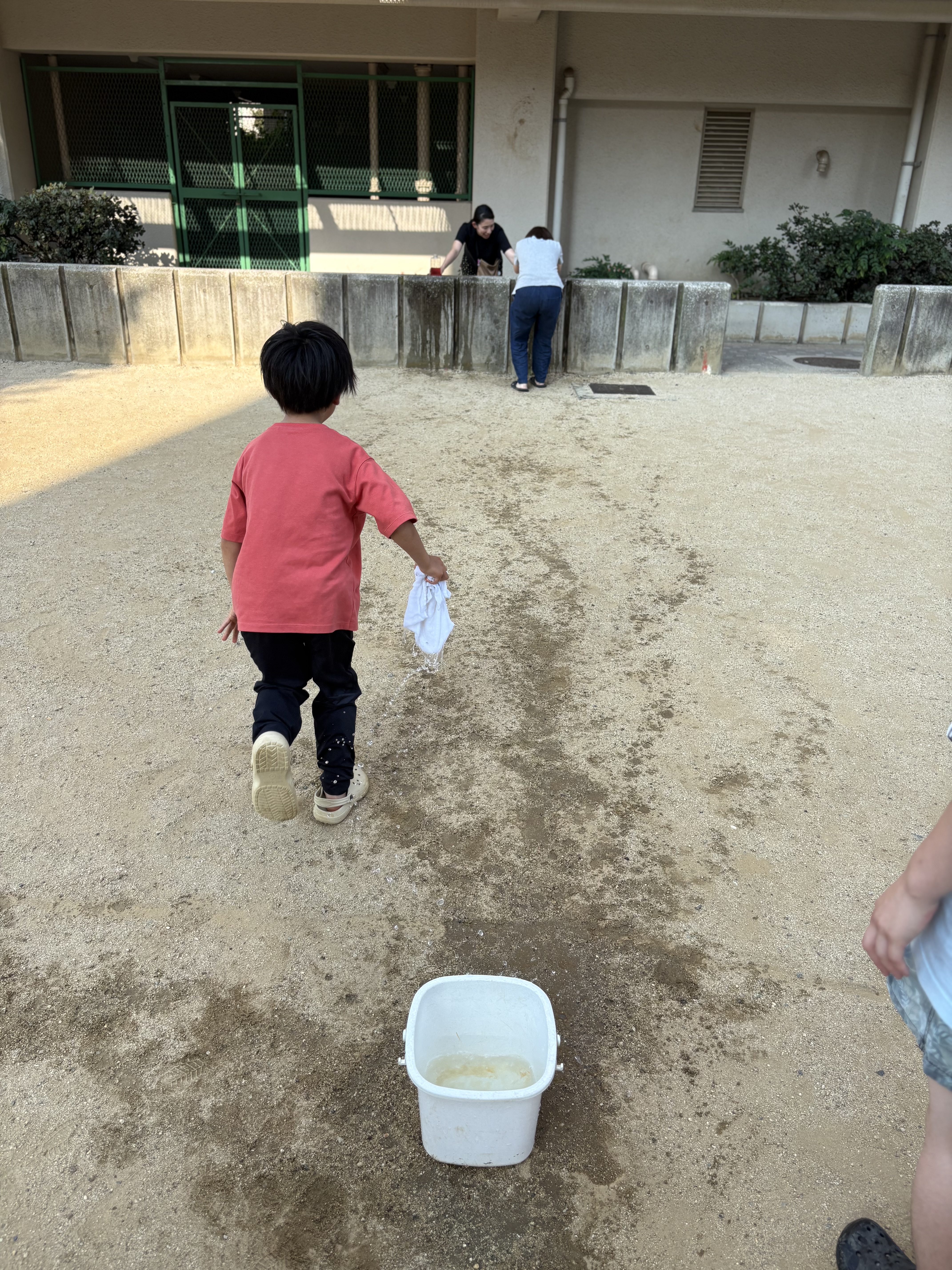「風を切って走る爽快感！涼しい夕方の公園を駆け抜けて」
夕方の心地よい風を受けながら、バトン代わりのタオルを持ってバケツからボトルへ一直線に走る子どもの凛々しい表情。