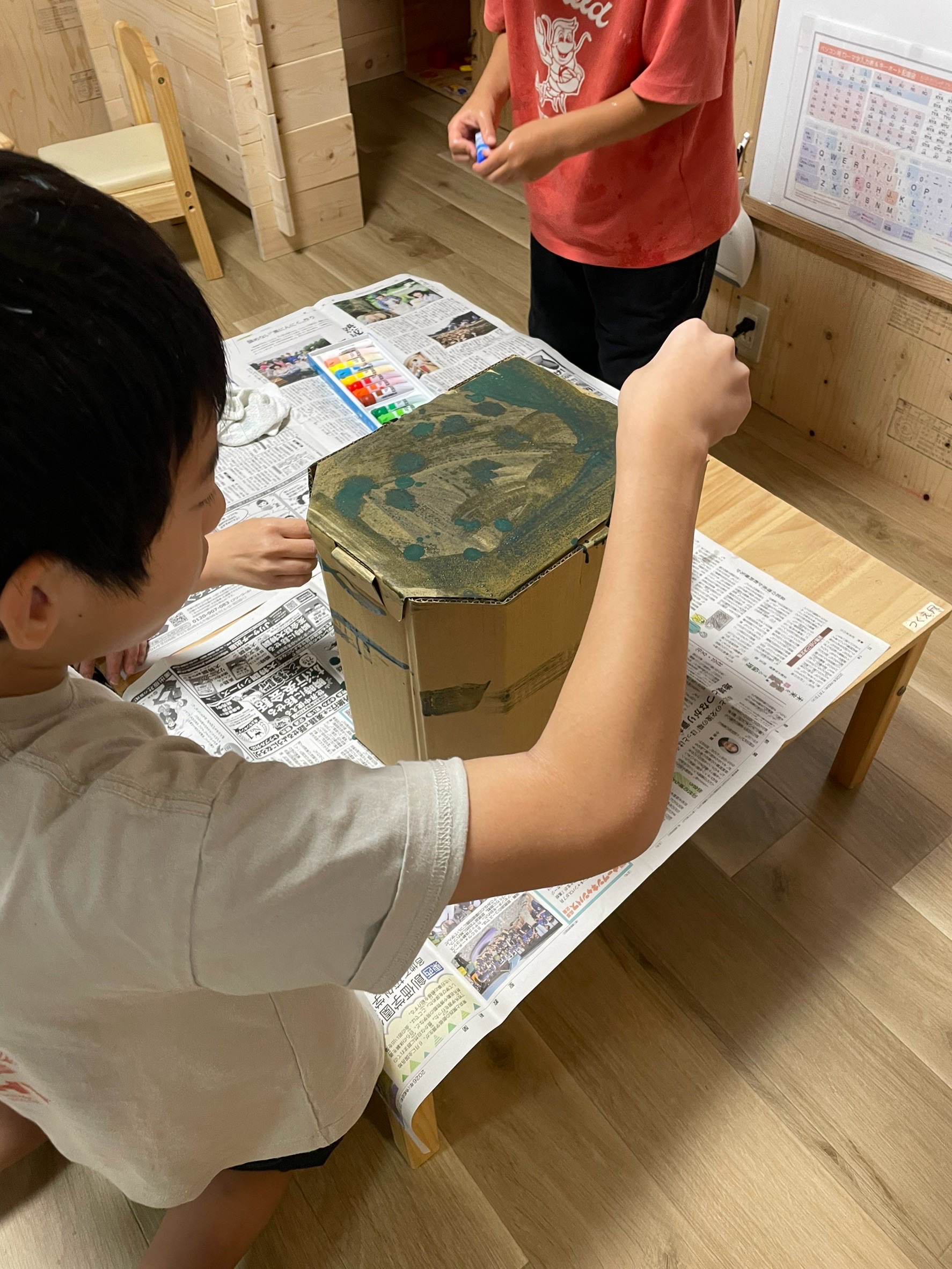 「『この大きな段ボールがキャンバス！？』ペイントスタート」
避難訓練で使った大きな組み立て式段ボール箱を囲み、刷毛や絵の具を用意して「さあ塗るぞ！」とワクワクしているオープニングショット。