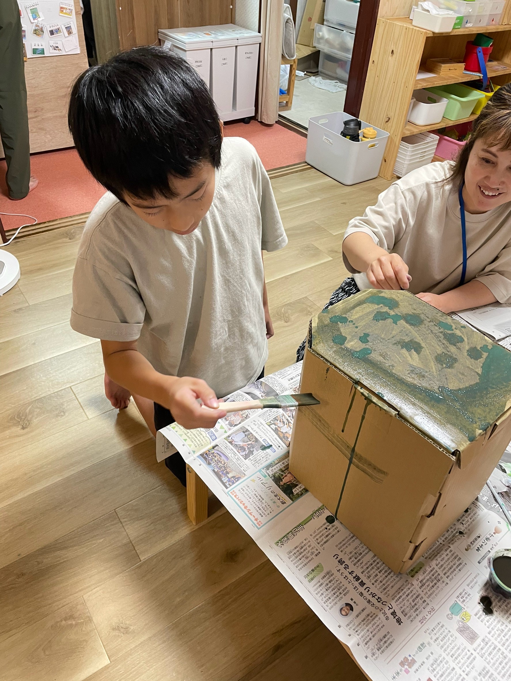 「『色を重ねたらカッコよくなった！』段ボールにしみ込む発見」
絵の具が段ボールにしみ込んで変化する様子を間近でのぞき込み、「見て見て！」と色重ねを楽しんでいるカット。