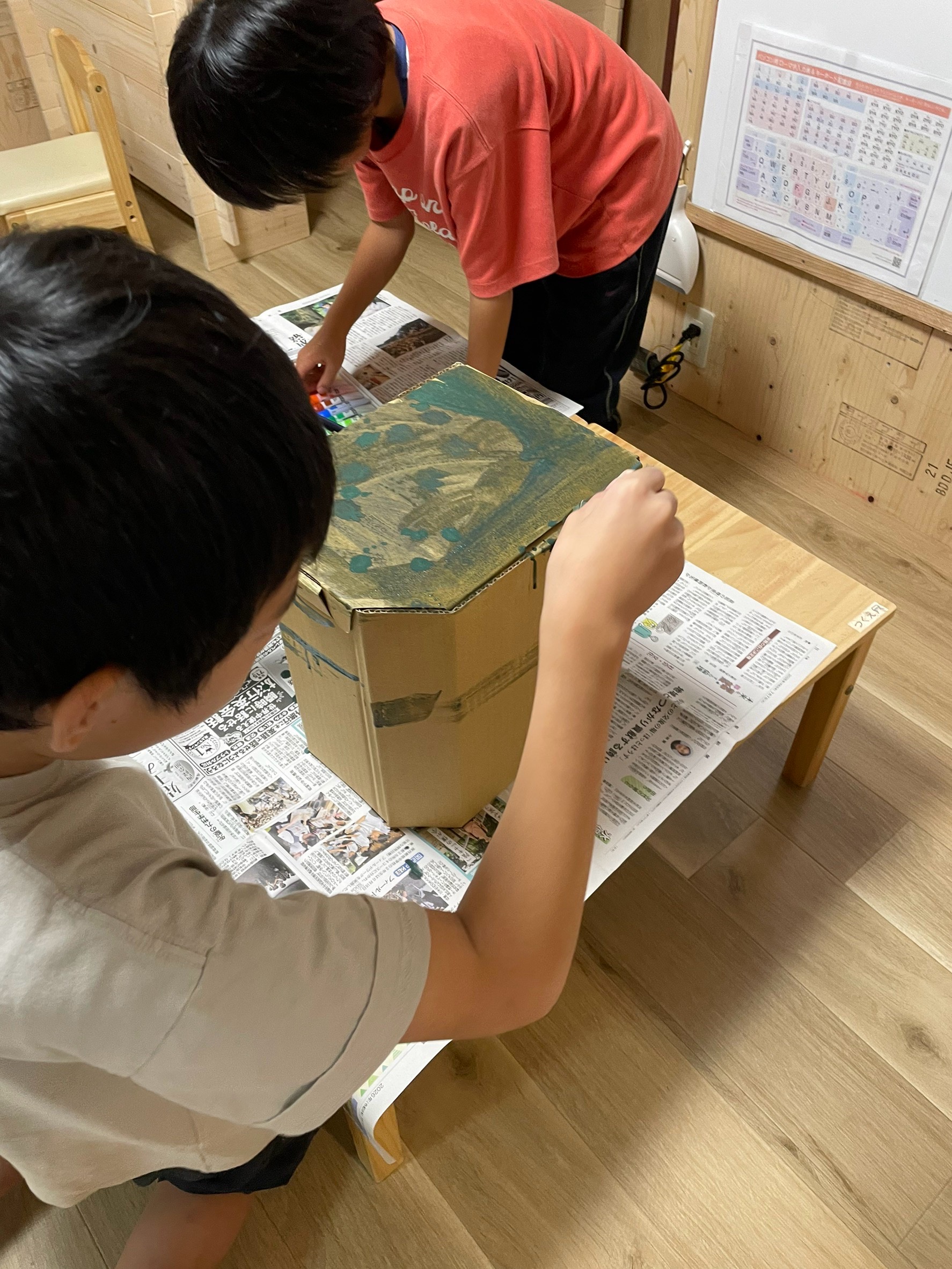 「『すーっと重なる色がおもしろい！』ダイナミックな筆使い」
深緑や茶色、黒などの絵の具を刷毛にたっぷりつけて、段ボールの表面に力強く塗っている真剣な表情と手元。