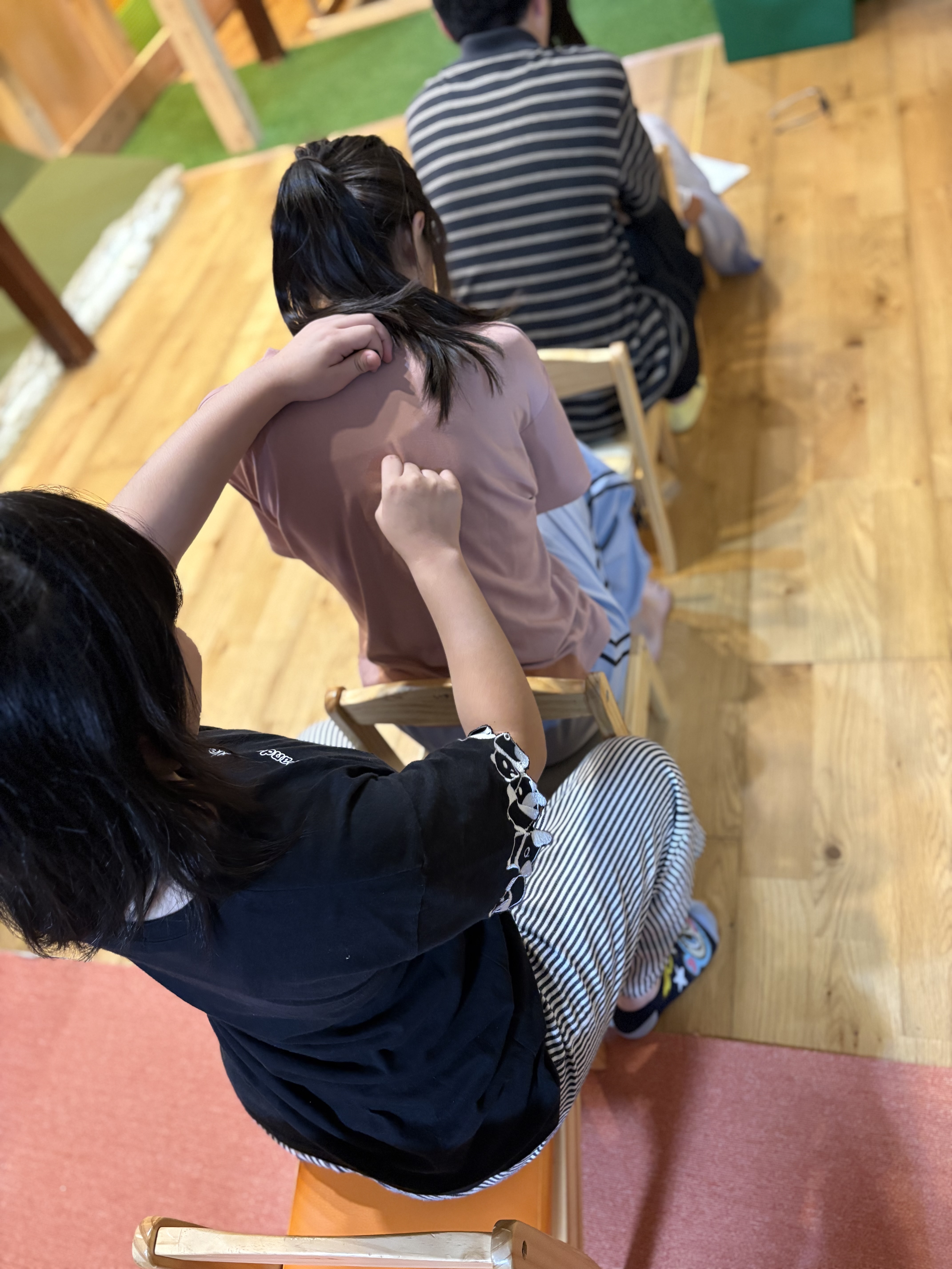 「『かきかき…そーっと指先でなぞる手元』お題を伝える瞬間」
後ろのお友達が、前のお友達の背中に指先で丁寧に文字や図形を描き込む手元のアップショット。